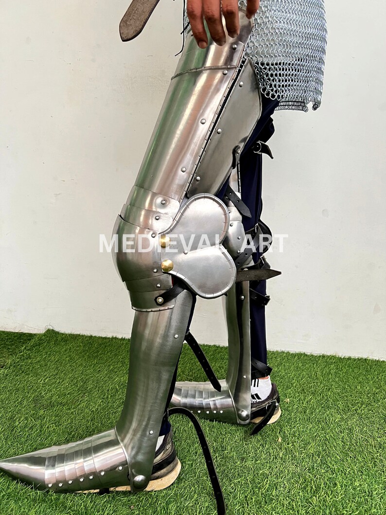 May include: A pair of silver metal leg armor with a pointed toe, knee pads, and straps. The armor is worn over blue pants and white socks.