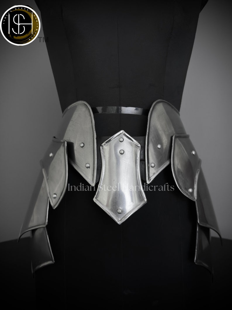 May include: A silver metal waist chain with a pointed center piece and multiple plates. The chain is on a black mannequin.