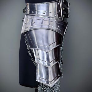 K&ouml;nnte beinhalten: Ein silberfarbenes Beinpanzerteil aus Metall mit Nietakzenten und einem Kettenrock. Die R&uuml;stung ist mit Lederriemen und Schnallen an einer schwarzen Form befestigt. Das Design erinnert an mittelalterliche oder Fantasy-Kleidung.