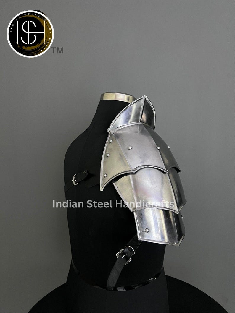 May include: A silver metal shoulder armour with a pointed top and multiple plates. The armour is attached to a black mannequin with a leather strap.