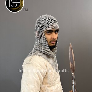May include: A silver chainmail coif with a hood, worn by a person holding a long, pointed, metal spear. The person is wearing a white quilted jacket. The image includes the text "Indian Steel Handcrafts".