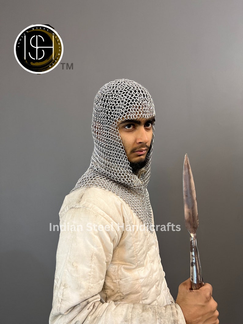 May include: A silver chainmail coif with a hood, worn by a person holding a long, pointed, metal spear. The person is wearing a white quilted jacket. The image includes the text "Indian Steel Handcrafts".