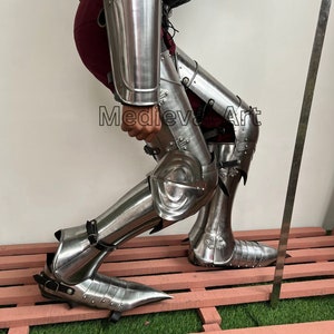 May include: A pair of silver metal leg armor with pointed shoes. The armor is worn by a person with a red skirt and a dark jacket. The text "Medieval Art" is visible on the armor.