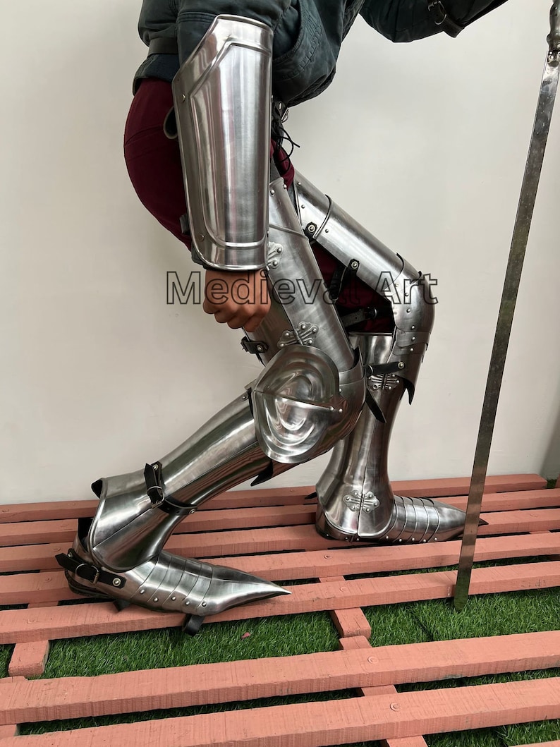 May include: A pair of silver metal leg armor with pointed shoes. The armor is worn by a person with a red skirt and a dark jacket. The text "Medieval Art" is visible on the armor.