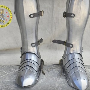 May include: A pair of silver metal shin guards with straps and buckles. The shin guards are made of metal plates that are joined together. The shin guards are worn on the legs and extend from the knee to the ankle.