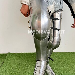 May include: A pair of silver metal medieval knight leg armor with pointed toes. The armor is made of metal plates and has a shiny finish. The armor is worn on a person's legs and is secured with straps.