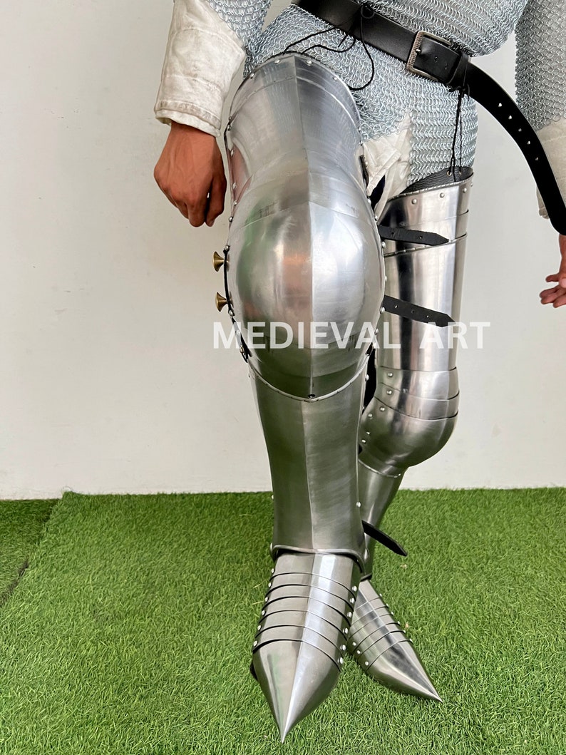 May include: A pair of silver metal medieval knight leg armor with pointed toes. The armor is made of metal plates and has a shiny finish. The armor is worn on a person's legs and is secured with straps.