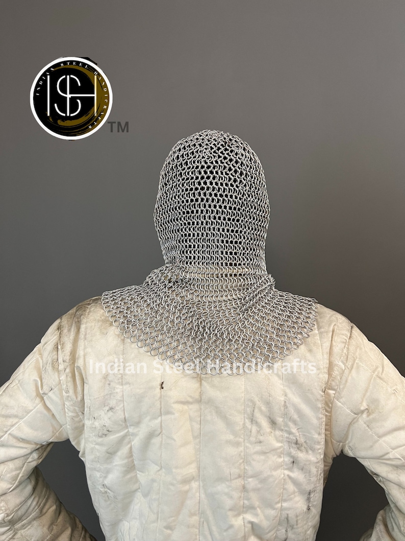 May include: A silver chainmail coif with a hood, worn over a white padded jacket. The coif is made of interlocking metal rings and covers the head and neck. The jacket has a quilted design and is worn for protection.
