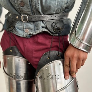 May include: A pair of silver metal leg armor with black leather straps and buckles. The armor is worn over a pair of red pants. The text "Medieval Art" is visible on the armor.
