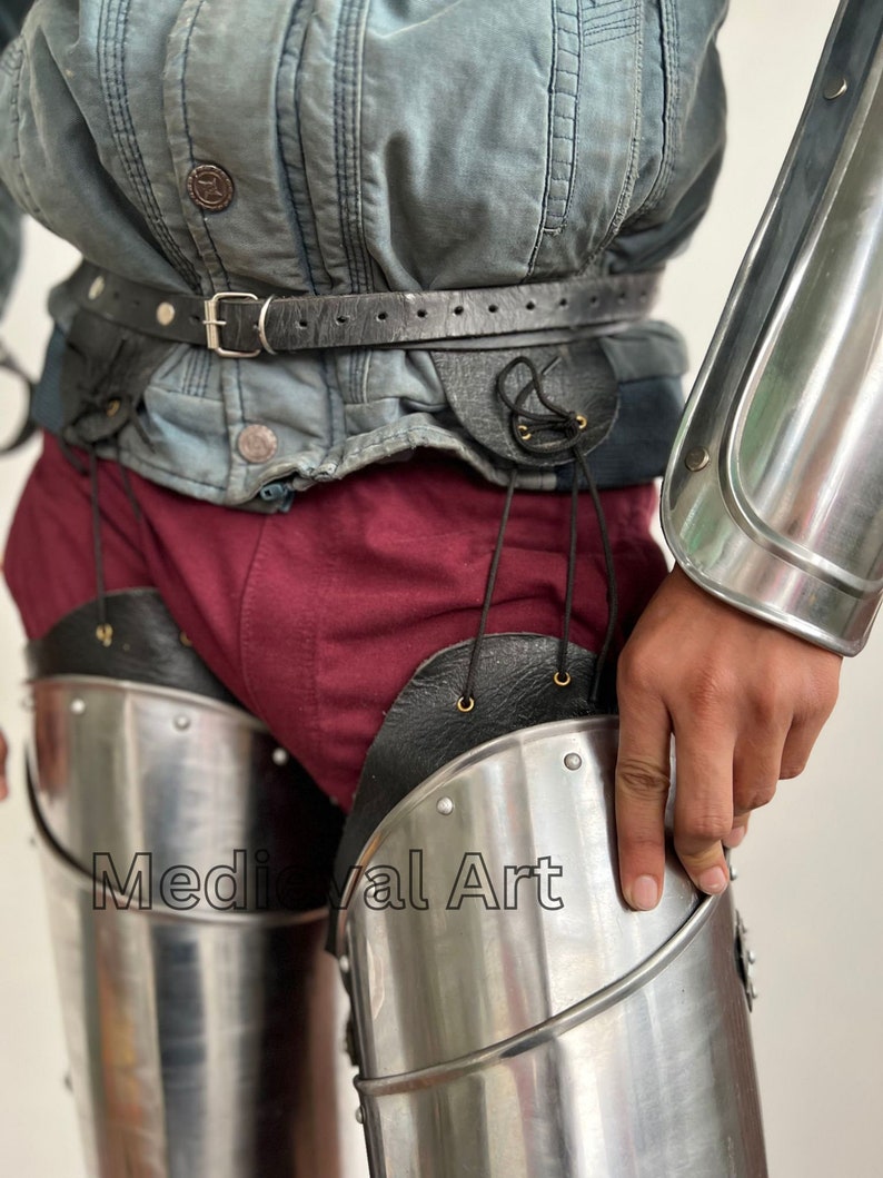 May include: A pair of silver metal leg armor with black leather straps and buckles. The armor is worn over a pair of red pants. The text "Medieval Art" is visible on the armor.