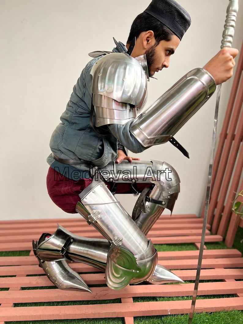 May include: A person wearing medieval armor, including shin guards, knee pads, and arm guards, is kneeling on a wooden platform. The armor is made of polished metal and has a shiny finish. The person is holding a long, thin sword in their right hand.
