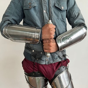 May include: A pair of silver metal arm guards and a pair of silver metal leg guards. The arm guards are worn on the forearms and the leg guards are worn on the thighs. The guards are made of metal plates that are connected by leather straps.