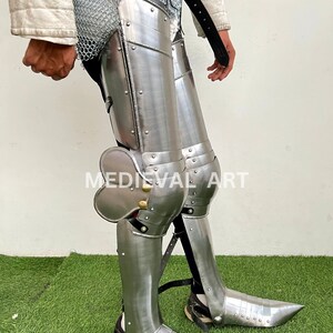 May include: A pair of silver metal leg armor with knee pads and pointed shoes. The armor is made of metal plates and has rivets and straps. The armor is worn over chainmail.