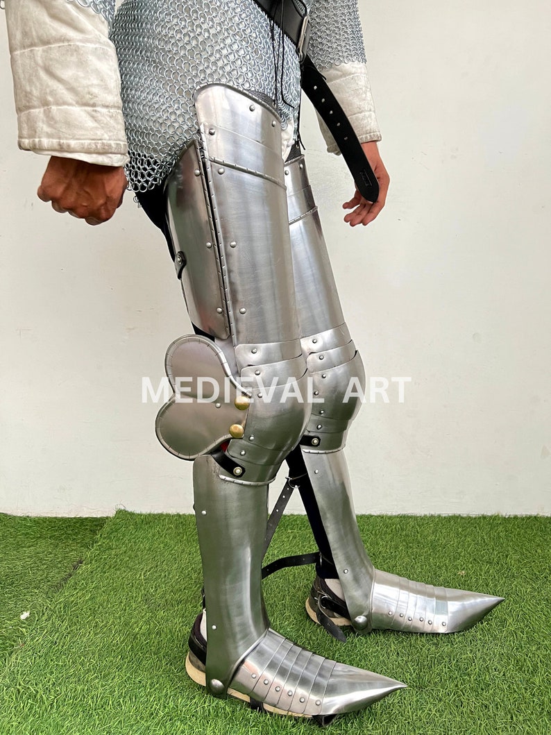 May include: A pair of silver metal leg armor with knee pads and pointed shoes. The armor is made of metal plates and has rivets and straps. The armor is worn over chainmail.