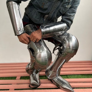 May include: A set of medieval-style armor for the legs and arms. The armor is made of metal and has a silver finish. The armor is worn by a person kneeling on a wooden platform.