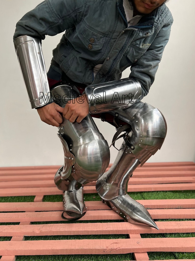 May include: A set of medieval-style armor for the legs and arms. The armor is made of metal and has a silver finish. The armor is worn by a person kneeling on a wooden platform.