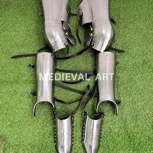 May include: A pair of silver medieval leg armor with black leather straps. The armor includes shin guards, knee guards, and greaves. The armor is on a green artificial grass background.