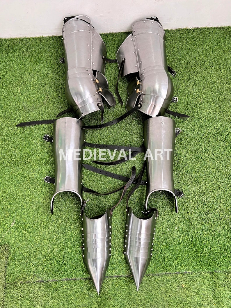 May include: A pair of silver medieval leg armor with black leather straps. The armor includes shin guards, knee guards, and greaves. The armor is on a green artificial grass background.