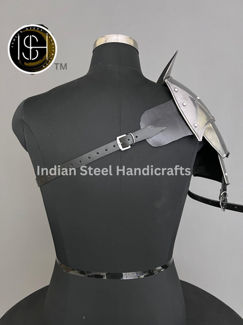 May include: A silver and black shoulder armour with a pointed top and a leather strap. The armour is worn on a black mannequin. The text "Indian Steel Handicrafts" is visible below the armour.