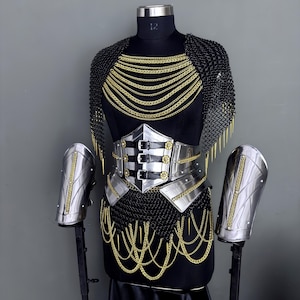 May include: A medieval-style costume displayed on a mannequin. The ensemble includes a black chainmail top, a silver and black corset with buckles, and matching arm guards. Gold chains and accents add detail.