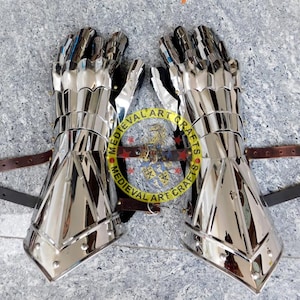May include: A pair of silver plated medieval gauntlets with leather straps. The gauntlets are made of metal plates and have a pointed design. The gauntlets are worn on both hands and are secured with straps.