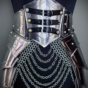 K&ouml;nnte beinhalten: Ein silberfarbener Metallkorsett mit schwarzen Lederriemen und Schnallen. Das Korsett hat genietete Akzente und einen Kettenrock. Das Design wird auf einer schwarzen Schaufensterpuppe pr&auml;sentiert.