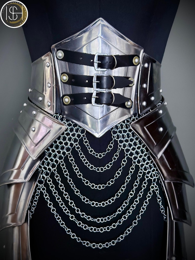K&ouml;nnte beinhalten: Ein silberfarbener Metallkorsett mit schwarzen Lederriemen und Schnallen. Das Korsett hat genietete Akzente und einen Kettenrock. Das Design wird auf einer schwarzen Schaufensterpuppe pr&auml;sentiert.