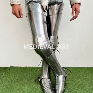 May include: A pair of silver-colored metal leg armor with pointed toes. The armor is worn over chainmail and has a decorative design of rivets.