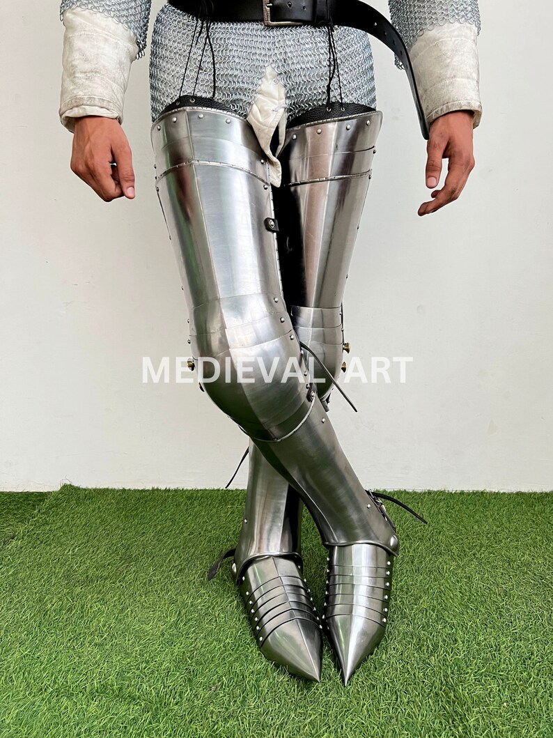 May include: A pair of silver-colored metal leg armor with pointed toes. The armor is worn over chainmail and has a decorative design of rivets.