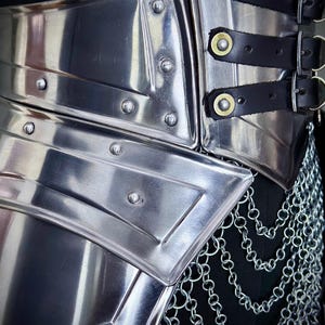 K&ouml;nnte beinhalten: Nahaufnahme einer polierten Metallr&uuml;stung mit genieteten Details und schwarzen Lederriemen. Die R&uuml;stung verf&uuml;gt &uuml;ber einen Kettenrock und ist zum Schutz des Torsos konzipiert. Das Metall hat eine reflektierende Oberfl&auml;che und erinnert an mittelalterliche R&uuml;stungen.