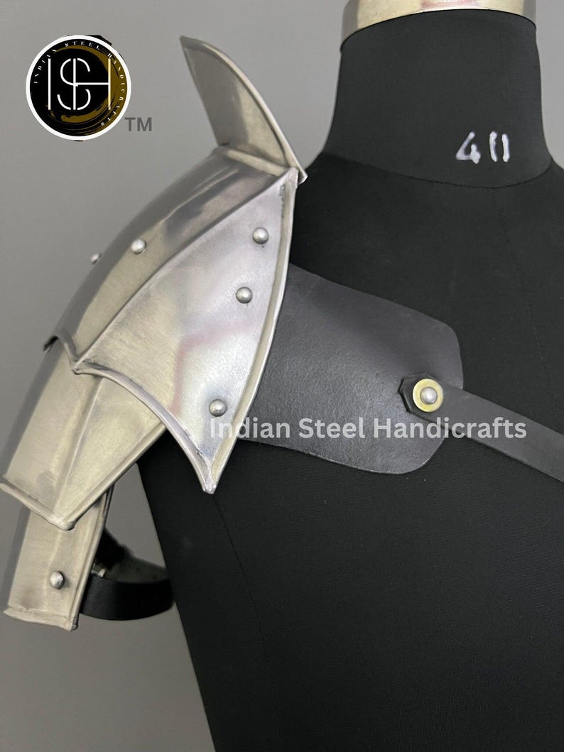 May include: Silver metal shoulder armour with a black leather strap and a gold buckle. The armour is worn on a black mannequin. The text "Indian Steel Handicrafts" is visible on the strap.