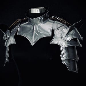 May include: A silver-coloured, medieval-style armour set, including a gorget and pauldrons, is displayed against a black background. The armour features intricate designs and leather straps. The shoulder armour is layered and articulated.