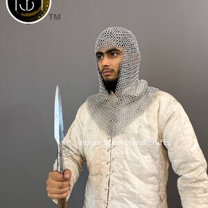 May include: A person wearing a white padded jacket and a silver chainmail coif, holding a long, silver-tipped spear. The text "Indian Steel Handicrafts" is visible on the jacket.