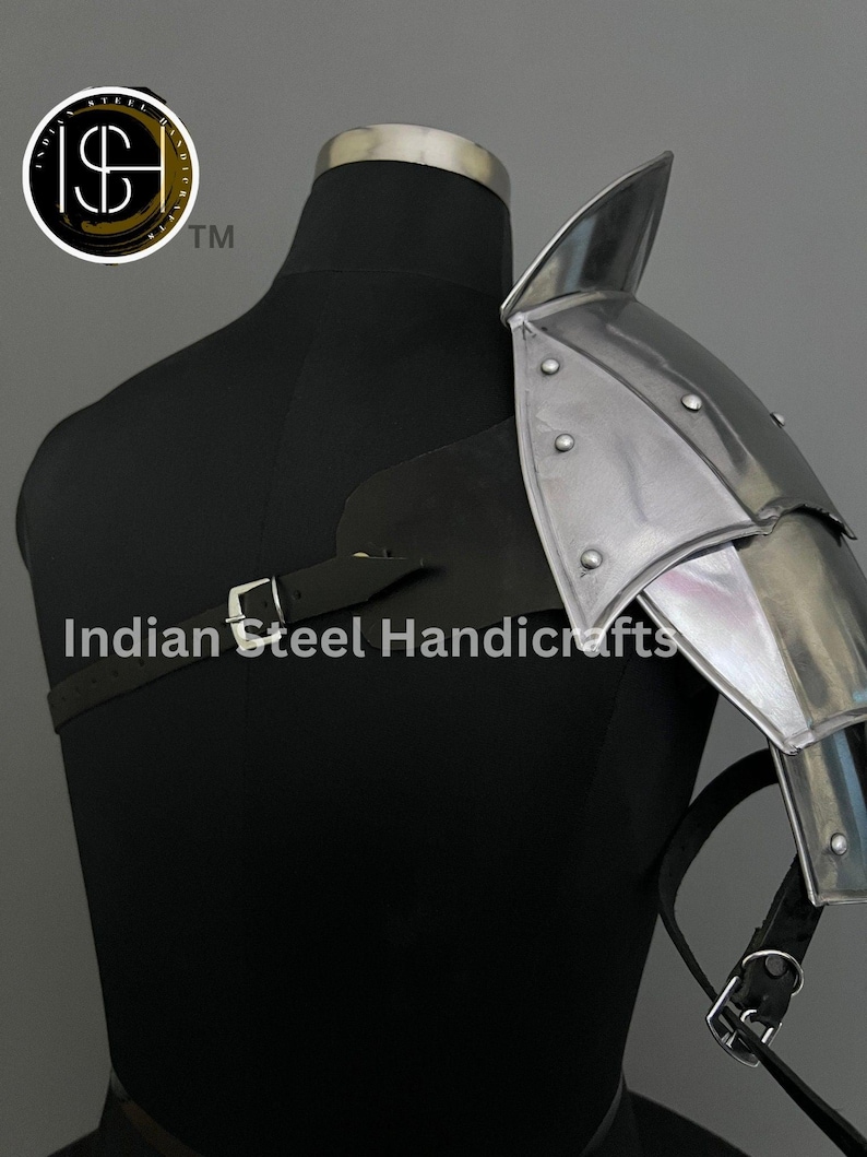 May include: A silver metal shoulder armour with a pointed top and a leather strap. The armour is attached to a black mannequin.