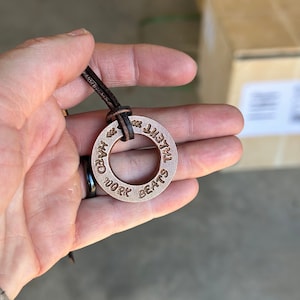 May include: A copper-coloured necklace with a round pendant that reads "Hard Work Beats Talent". The pendant is attached to a brown leather cord.