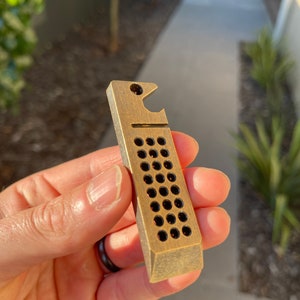 May include: A brass bottle opener with a hole pattern and a bottle opener on the top.