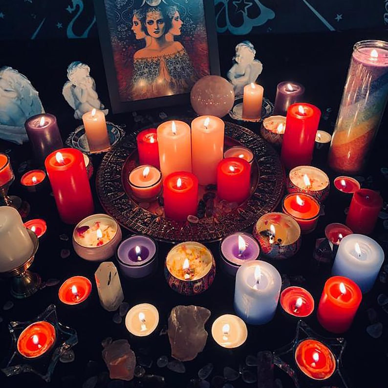 May include: A variety of candles in different colors and sizes are arranged in a circle on a black surface. There is a small painting of a woman with three faces in the background. The candles are surrounded by crystals and other decorative items.