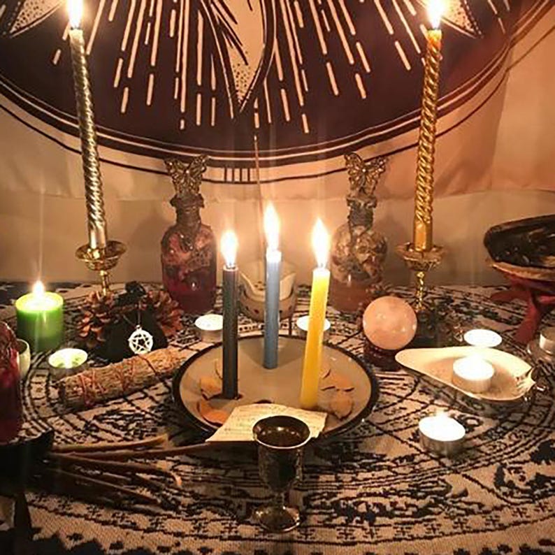 Pu&ograve; includere: Una tavola apparecchiata per un rituale o un incantesimo con candele, cristalli e altri oggetti magici. Ci sono quattro candele, una nera, una blu, una gialla e una verde. C'&egrave; una bottiglia di vetro trasparente con un liquido rosso e una sfera di cristallo. Il tavolo &egrave; coperto da una tovaglia a motivi.