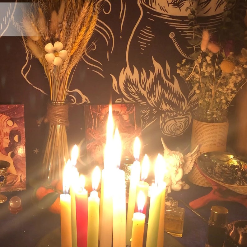 May include: A group of lit candles in various colors, arranged in a circle on a dark surface. The candles are surrounded by dried flowers, a small white figurine, and other decorative items.