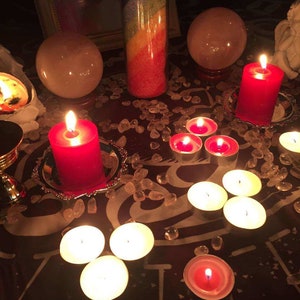 May include: A red candle sits on a silver plate surrounded by small clear crystals. There are several white tea lights and a larger red candle on a table with a clear glass jar filled with colorful wax.