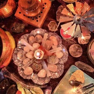 May include: A crystal grid featuring rose quartz crystals, selenite sticks, and a candle at its centre. The grid is surrounded by other crystals, including a clear quartz point, a clear quartz cluster, and a clear quartz sphere. The grid is set on a dark red surface with a star-like pattern.
