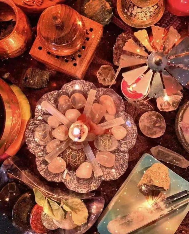 May include: A crystal grid featuring rose quartz crystals, selenite sticks, and a candle at its centre. The grid is surrounded by other crystals, including a clear quartz point, a clear quartz cluster, and a clear quartz sphere. The grid is set on a dark red surface with a star-like pattern.