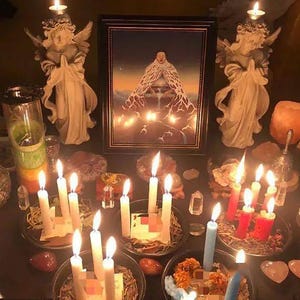 K&ouml;nnte beinhalten: Ein spiritueller Altar mit brennenden Kerzen, Engelsstatuen und einem gerahmten Bild. Die Kerzen sind wei&szlig;, rot und blau und in kleinen Schalen angeordnet. Das Bild zeigt eine Figur in einem flie&szlig;enden wei&szlig;en Gewand. Die Szene ist schwach beleuchtet.
