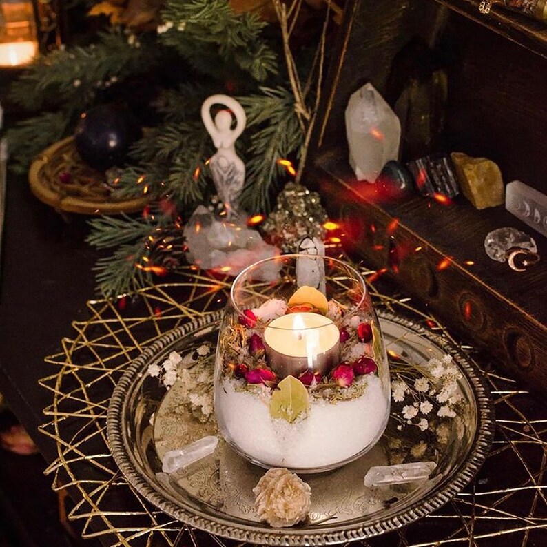 May include: A glass jar filled with white sand, dried flowers, and a small white candle. The jar is sitting on a silver tray with a gold metal base. There are several crystals and other decorative items around the jar.