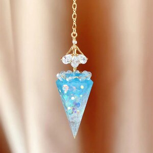 May include: A blue crystal pendulum with gold accents and a clear crystal top. The pendulum is hanging from a gold chain.