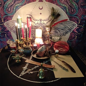 Pu&ograve; includere: Un telo da altare nero con un disegno di pentagramma bianco, circondato da vari oggetti tra cui candele, un calice, una piuma, una campana, una lampada del genio, una bacchetta di legno, una piccola ciotola, una bottiglia di olio, un mazzo di salvia e un pezzo di carta. L'altare &egrave; allestito davanti a un arazzo con un disegno di farfalla.