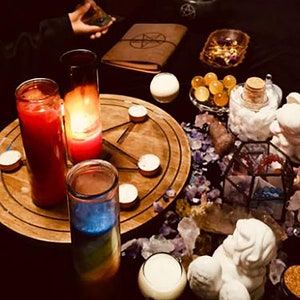 May include: A wooden table with a pentagram design and five small candles. There are two tall glass candles, one red and one with a blue and rainbow layered wax. The table is surrounded by crystals, a small white angel figurine, and a glass jar with white crystals.