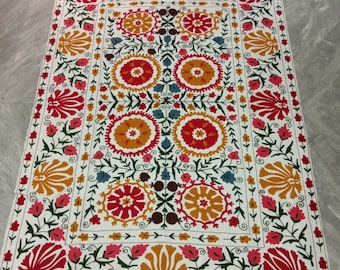 May include: A white cotton rug with a colorful floral pattern. The rug features a central square with a repeating design of red, yellow, and blue flowers. The border of the rug is decorated with a similar floral pattern.