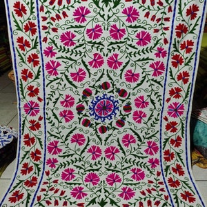 May include: White fabric with a floral embroidery design. The embroidery features pink, red, green, and blue flowers. The design is symmetrical and has a central motif.
