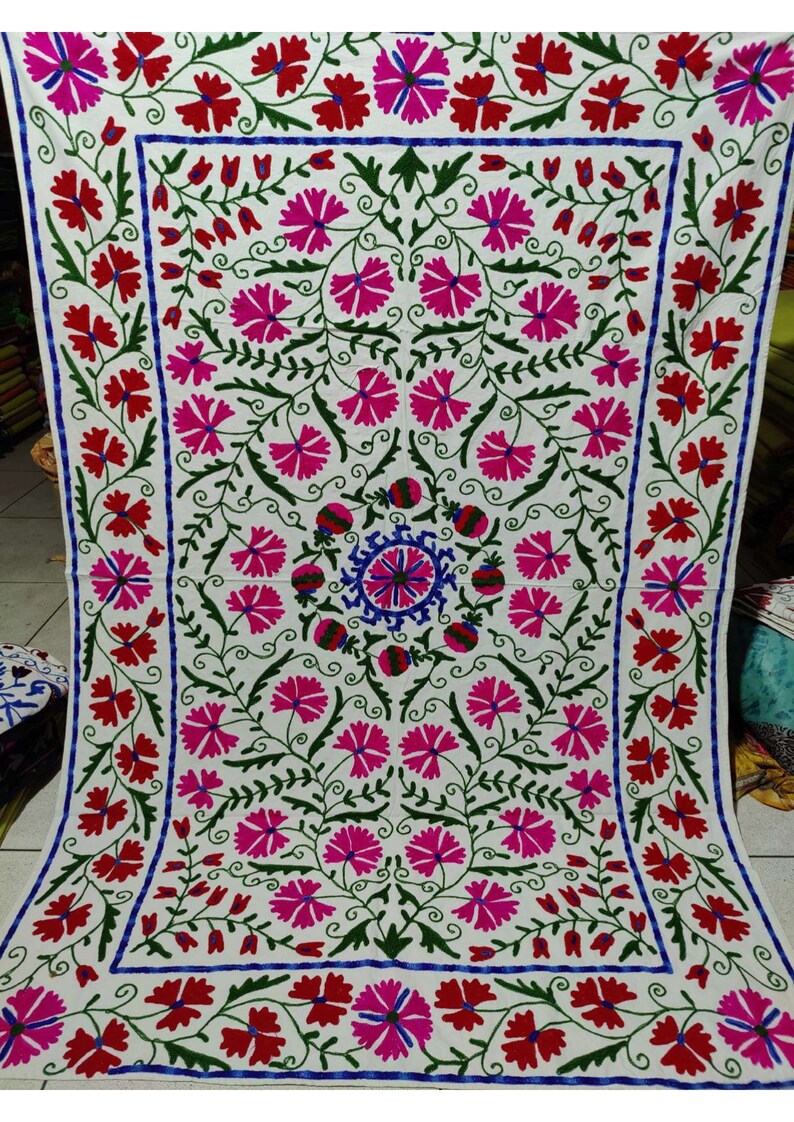 May include: White fabric with a floral embroidery design. The embroidery features pink, red, green, and blue flowers. The design is symmetrical and has a central motif.